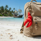 Beige backpack with a red lobster-claw-shaped keychain on a sandy beach with a ship and palm trees in the background.